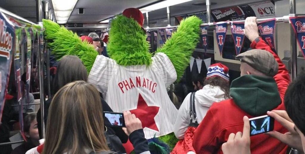 The Phillie Phanatic rides a subway full of Phillies fans, holding onto two overhead railings. He is visible from the back.