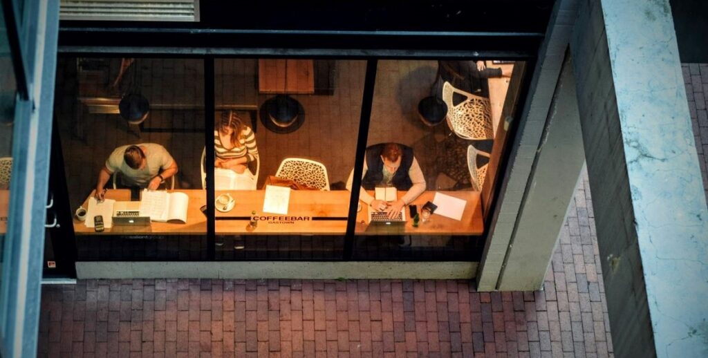 A man, a woman, and another man are seen through the window of a cafe working