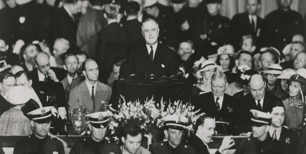 Black and white image shows U.S> President Franklin D. Roosevelt in 1936 at a podium before microphones, elevated above a crowd of white men and women, some in uniform, at the 1936 Democratic National Convention in Philadelphia, PA.