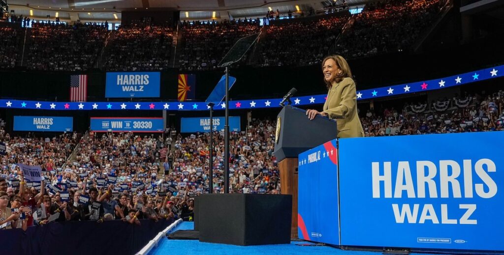 Democratic presidential candidate and current Vice President Kamala Harris, a biracial woman of Indian and Jamaican descent, stands at a podium in an arena. Alongside her is a large blue and white sign that says "HARRIS WALZ."