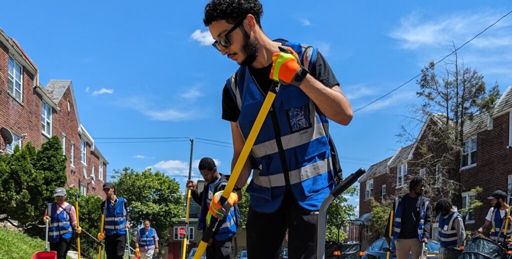 A bearded man with curly black hair and dark sunglasses wears a reflective blue vest over a black t-shirt and pants, and bright gloves while sweeping a city street where more people in similar dress are doing the same work.