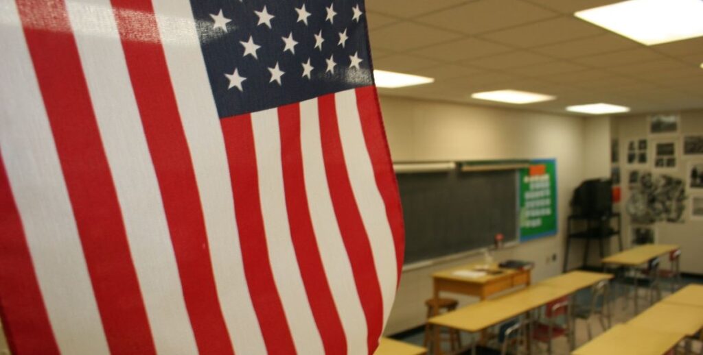 An American flag is partially visible on the left, behind it is an empty classroom with a green chalkboard