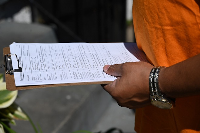 A brown hand with a wristwatch holds a clipboard holding a typed list of names and addresses of formerly incarcerated Philadelphians who are not registered to vote. The person holding the clipboard is wearing an orange t-shirt.