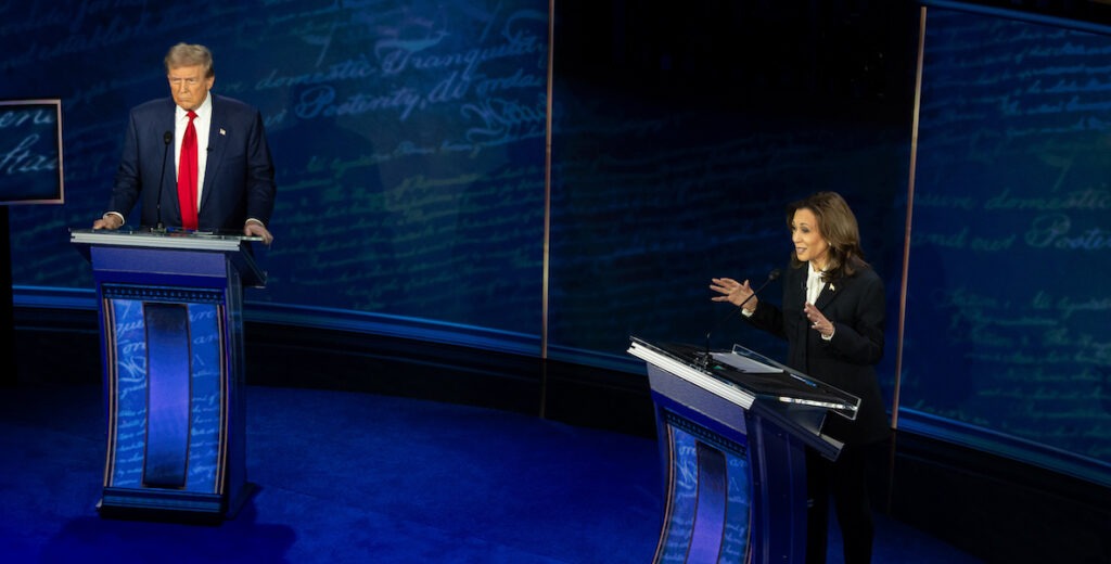 Former President Donald Trump and Vice President Kamala Harris debating at the National Constitution Center in Philadelphia on September 10, 2024. Donald Trump is a 78-year-old White man with blonde hair, wearing a navy blue suit jacket, white collared shirt, red tie and flag pin. He appears to be speaking, with his right hand somewhat raised. Kamala Harris is a 59-year-old Indian-African-American woman with past-shoulder length dark hair wearing pearl earrings, a black suit jacket, white blouse and flag pin. She is speaking and gesturing with both hands. Behind them are words, in script, from the U.S. Constitution.