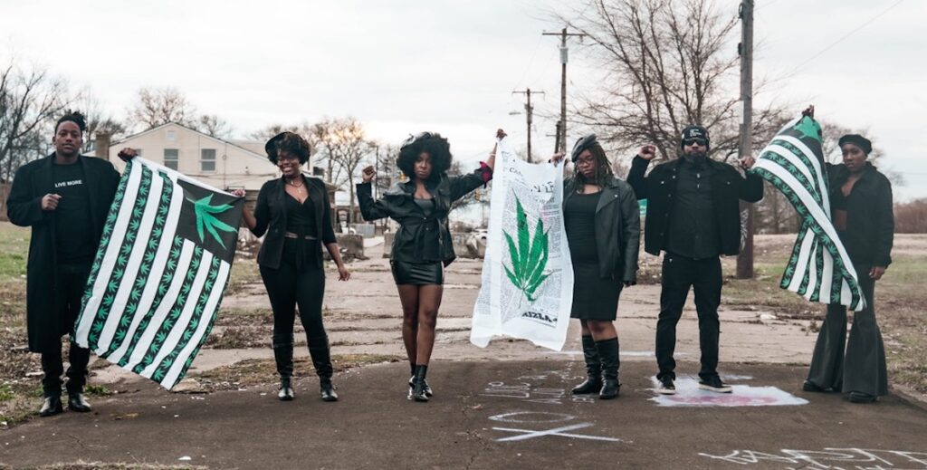 Kristal Bush (center) and other pro-recreational marijuana activists represent Black Cannabis Week by standing outside, wearing black, holding flags with marijuana leaves on them. Everyone in the line is stylish and African American.