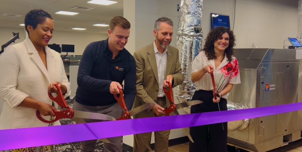 Carbon Reform capsule ribbon cutting at BGE. Left to right: Faith Davis (BGE, Lead Exelon's Climate Investment Fund), Nick Martin (Carbon Reform, COO & Co-Founder), Alexander Núñez (BGE, SVP, Governmental, Regulatory, and External Affairs), Jo Norris (Carbon Reform, CEO &Co-Founder). Four people hold giant scissors to cut a purple ribbon while standing beside a large silver machine with silver ducts sticking out of it.