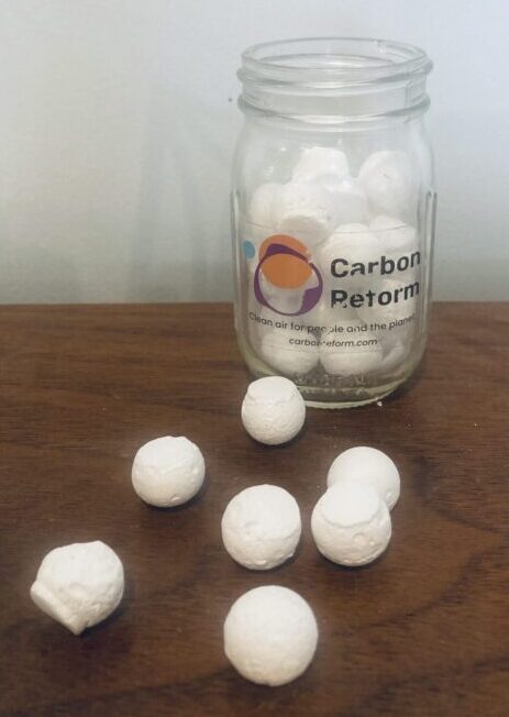 A glass jar bearing the logo of and words "Carbon Reform" holds small white orbs, these are carbon pellets produced by Carbon Reform's capture capsule. Seven additional round pellets rest on a wooden table.