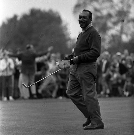 Pro golfer Charlie Sifford stands on a green and smiles. He has just won the 1969 Los Angeles Open. He is a Black man with short hair, holding a golf club, wearing a sweater and slacks. The photo is black and white. In the background spectators watch. Behind them are tress.