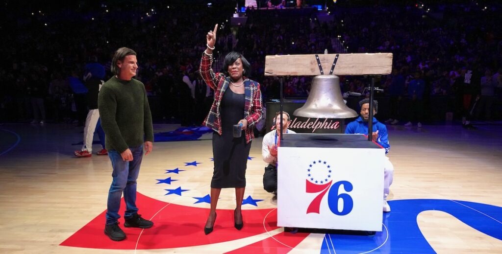 In the center of the 76ers basketball court of the Wells Fargo Center, atop a large 76ers logo, Philadelphia Mayor Cherelle Parker, a Black woman with ear-length black hair wearing a black dress, a plaid suit jacket, black heels, holding a rubber mallet in one hand and her pointer finger raised atop her head in a "one," stands alongside a replica Liberty Bell, which itself stands atop a white box with the 76ers logo. Next to her stands David Adelman, a co-owner of the 76ers, a white man with ear-length brown hair wearing a green sweater and jeans, smiling and standing facing the mayor. His arms hang at his sides. The background is dark, as this is the start of a home basketball game against the Houston Rockets at the Wells Fargo Center. Adelman and his co-owners have proposed leaving the Wells Fargo Center and building their own arena, 76 Place, on Market East on the edge of Philadelphia's Chinatown.