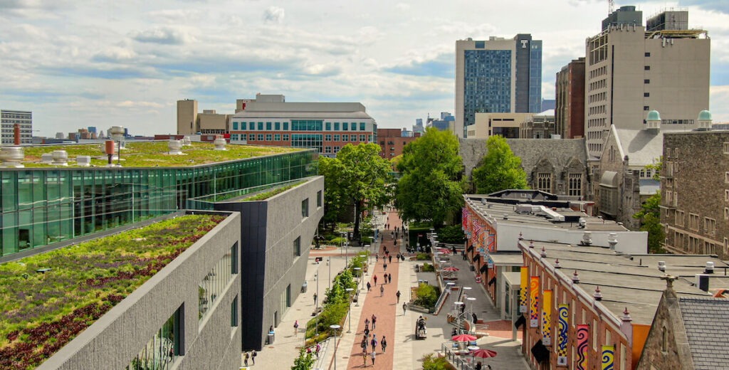 An aerial view of Temple University's Philadelphia campus features a brick walkway running between (left) two modern buildings with green roofs and (right) older brick buildings. Students are walking along the pathway. In the background is Temple University Hospital and more buildings.