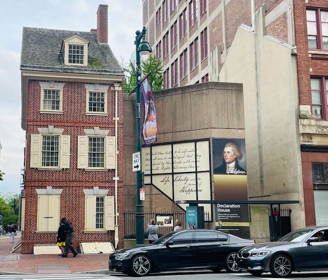 Declaration House before the installation, as viewed from Market Street. A slender brick townhouse stands at a corner of 7th and Market streets in Philadelphia. The paned windows have shutters, most open. Cars drive by. Next door is a concrete building bearing the image and cursive writing of Thomas Jefferson.