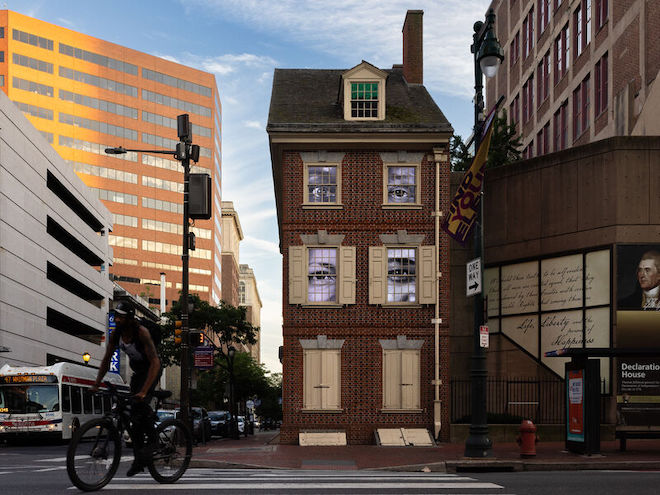 Sonya Clark's “The Descendants of Monticello,” a video installation of recordings of eyes of descendants of people Thomas Jefferson enslaved — installed at the brick replica Declaration House in Philadelphia where Jefferson wrote the Declaration of Independence, as viewed from the Market Street side of the Declaration House at Independence National Historical Park. Passing by the house are a cyclist (foreground) and a SEPTA bus. The structure stands alongside modern buildings.