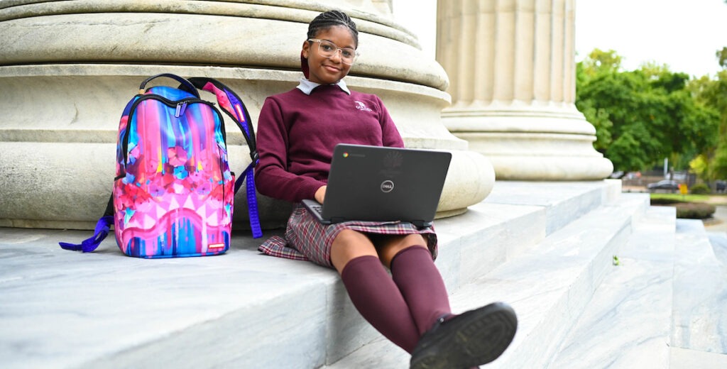 Faith Clanton, an African American sixth grader, wears her uniform from Girard College (burgundy sweater, plaid kilt, burgundy kneesocks, black shoes) along with clear glasses and braids pulled back while sitting on marble steps and leaning on a marble column. A Chromebook (laptop) sits in her lap. Her colorful backpack is at her side. She smiles slightly. Her ankles are crossed. Trees are in the background.