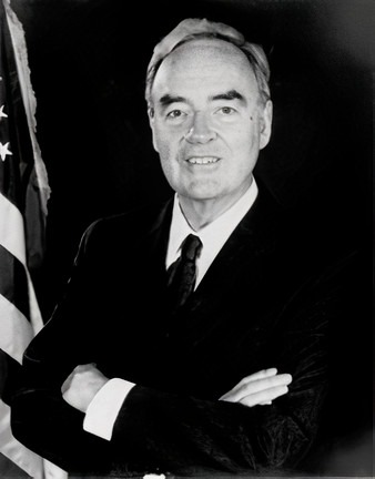 A black-and-white portrait of the U.S. Senator from Pennsylvania, Harris Wofford, a white man with greying hair and a slight smile stands beside an American flag and wears a dark suit and tie and white shirt. His arms are crossed.