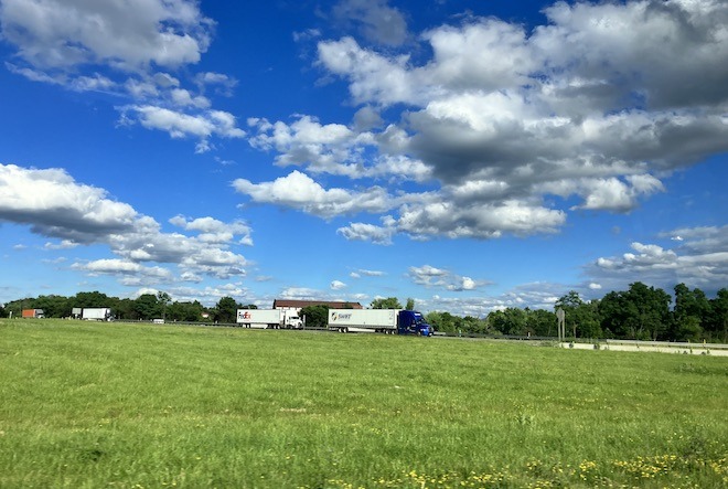 Between a verdant stretch of grass and bright blue, cloud-dotted sky runs a highway with 18-wheeler trucks in Harrisburg, PA. This is a scene from the Penn Empathy Lab.