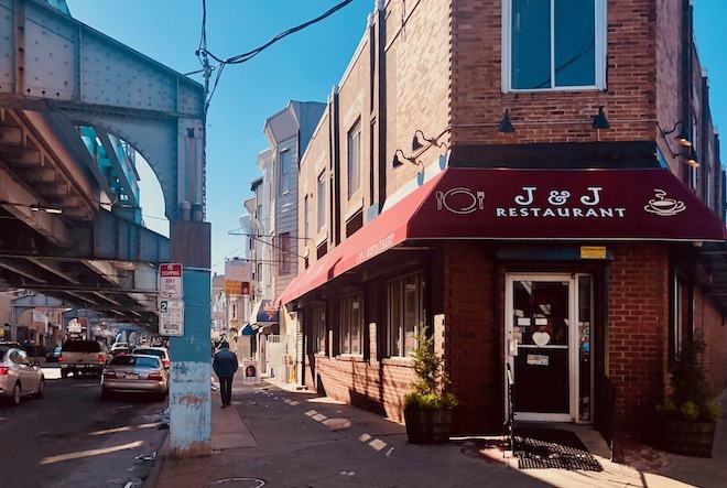 A red awning affixed to a brick building announces the entrance toJ & J Restaurant. On the other side of the sidewalk stands SEPTA's Market-Frankford El, in Kensington in Philadelphia.