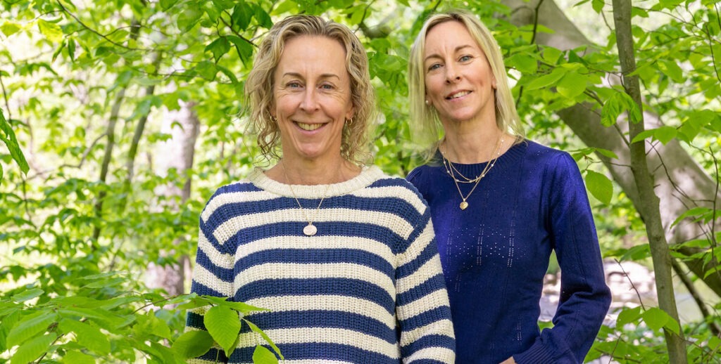 Jenn Asplundh (left, a white woman with curly blonde shoulder-length hair wearing gold jewelry and a striped sweater) and Katie Coffin (a white woman with shoulder-length straight hair wearing pearl earrings, gold necklaces and a blue knit top) stand side-by-side surrounded by trees. These are the sister-creators of the Kindred podcast.