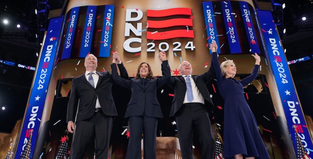 Left to right: Doug Emhoff, Vice President Kamala Harris, Minnesota Governor Tim Walz and Gwen Walz onstage at the Democratic National Convention, in Chicago, Illinois, Wednesday, August 22, 2024. All are wearing dark clothing and holding hands raised in the air while confetti floats down around them. Behind them are several signs that say "DNC 2024."