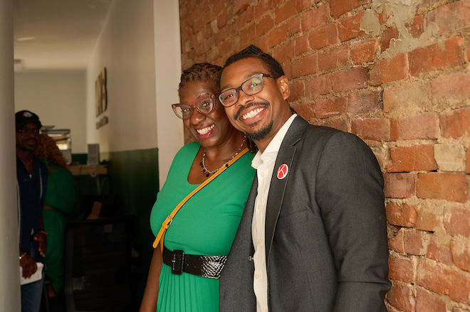 Two people, a woman and a man, stand with their backs to an exposed brick wall and smile. Both are African American and wear glasses. One is a woman in a green dress. The other is a man with a slight beard, grey suit jacket and white collared shirt.