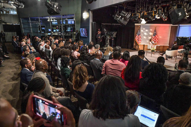 The room with Harris and members of the National Association of Black Journalists (NABJ). Photo by Jason M. Johnson for NABJ. The backs of a large group of Black journalists who are sitting facing a stage where Vice President and presidential candidate Kamala Harris is seated between two U.S. flags, facing three Black journalists, also seated.