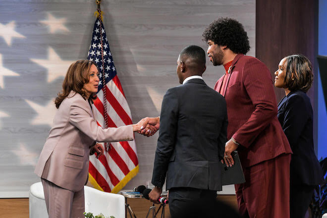 Kamala Harris, dressed in a khaki pantsuit, stands before a U.S. flag and shakes the hand of Eugene Daniels, a Black journalist in a burgundy suit, who is standing between Gerrn Keith Gaynor, a Black journalist in a dark suit, and Tanya Mosley, a Black journalist in a navy suit.