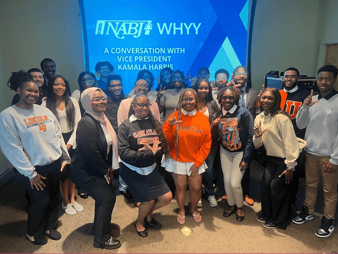 A group of Black students, many wearing college sweatshirts and sweaters, poses in front of a large screen that says "NABJ WHYY A CONVERSATION WITH VICE PRESIDENT KAMALA HARRIS."