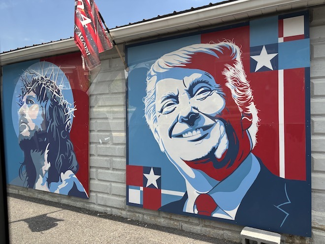 Two red, white and blue paintings of Jesus wearing a crown of thorns and a smiling Donald Trump (both from the shoulders up) hand along a white brick wall. Between them hangs a red and black striped flag. The paintings are in the style of Shepherd Fairey near Fulton County, PA.