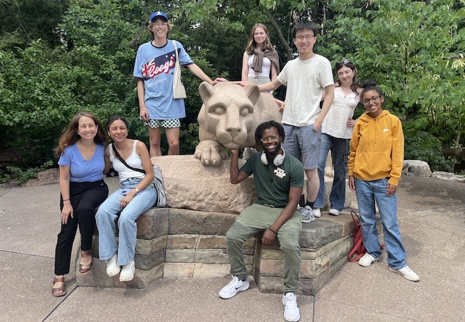 A diverse group of eight college age students in casual clothing pose around the Nittany Lion statue at Penn State.