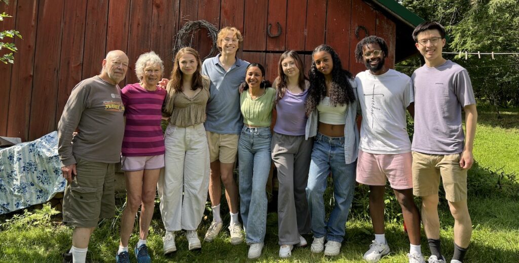 A diverse group of 7 college students, members of the Penn Empathy Lab, all wearing casual clothing stands alongside an older white couple, everyone arm in arm, in front of a faded red barn.