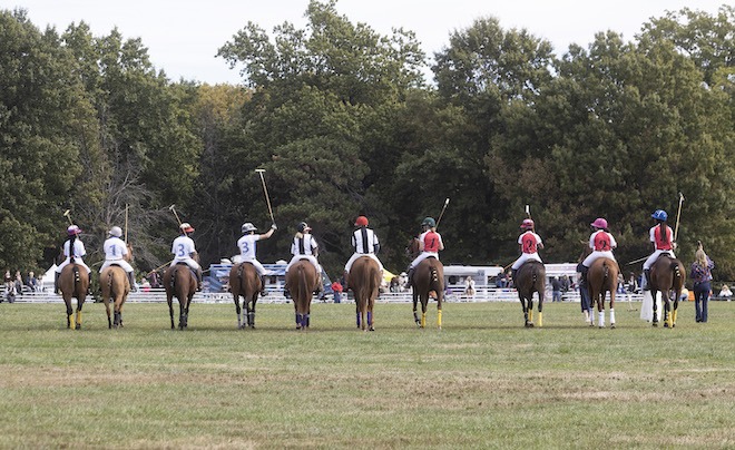 Riders dressed in polo gear ride away from the camera along a field at the Philadelphia Polo Classic.