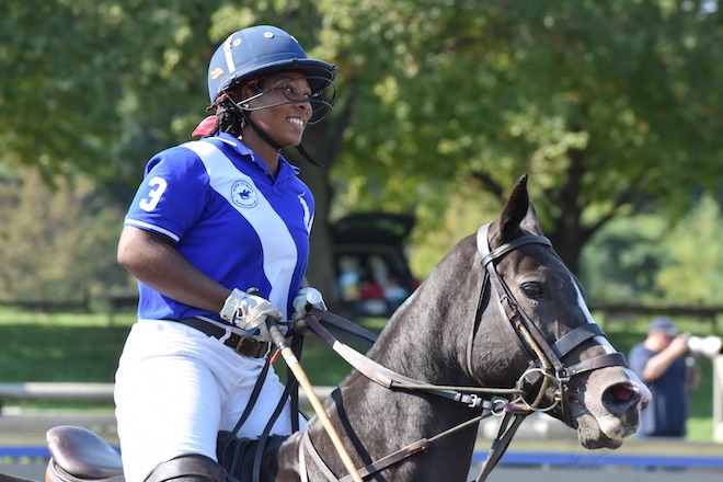 Shariah Harris, a Black athlete wearing white jodhpurs, a blue and a white polo, riding helmet and black books, rides a strong brown horse on a polo field at a previous Philadelphia Polo Classic.