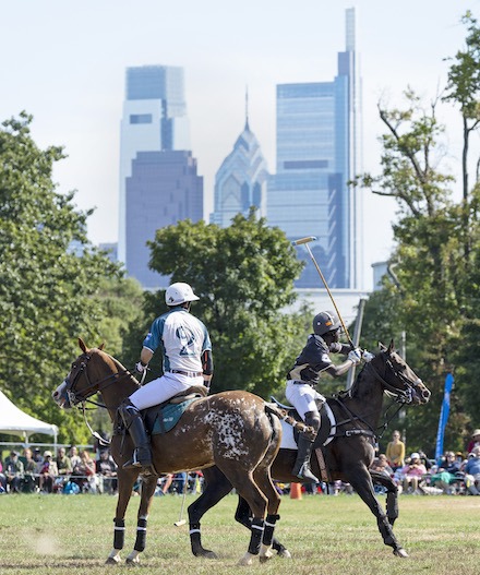 African American polo players play atop horses on a field with the skyline of Philadelphia in the background.