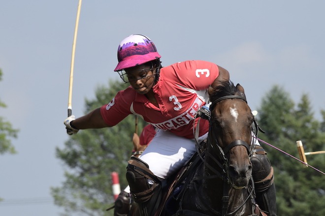 Shariah Harris, a Black high school-aged polo player raises a mallet while on horseback. She is wearing white jodhpurs, a red polo shirt with the number 3 and the word "Lancaster," and a bright pink and white helmet. Photo courtesy of Lezlie Hiner.