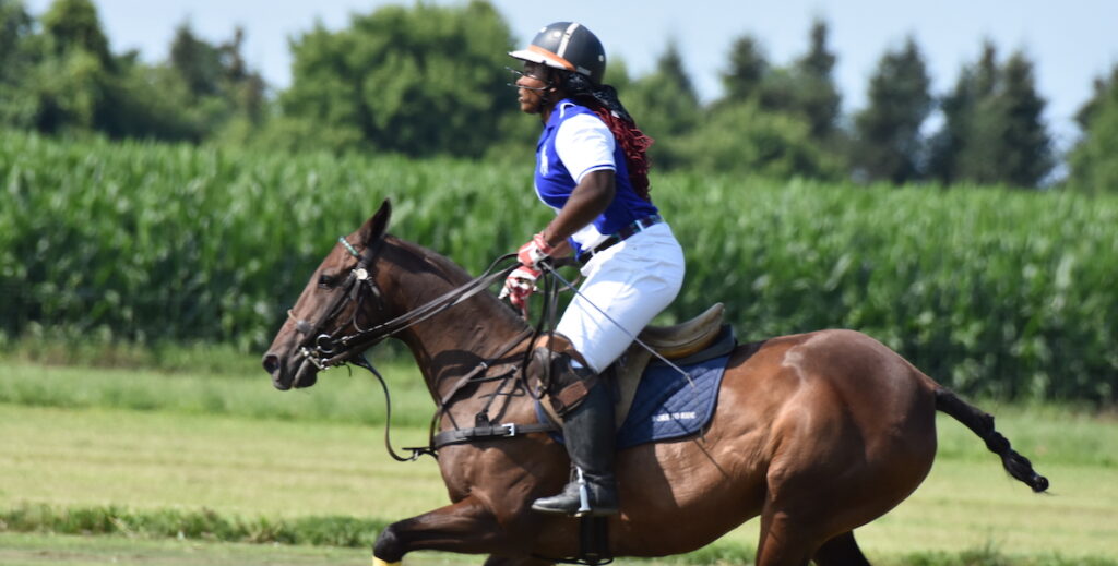 Shariah Harris, a Black athlete wearing white jodhpurs, a blue and a white polo, riding helmet and black books, rides a strong brown horse on a polo field at a previous Philadelphia Polo Classic.