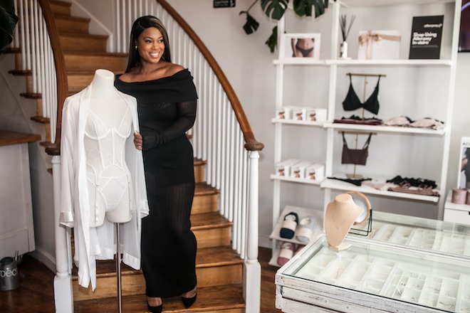 Shaw Lewis, owner of Expect Lace and Gild + Olive, is a Black woman with long dark straight hair with blonde streaks wearing a tight-fitting, off-the-shoulder, long-sleeved black dress. She stands before a curving wooden staircase alongside a headless mannequin wearing women's undergarments and a white robe. Lingerie is displayed to her left.