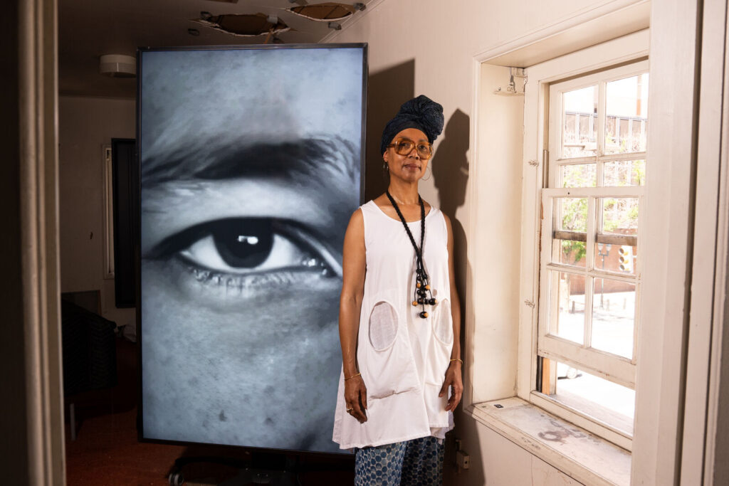 Sonya Clark, a Black woman wearing her hair wrapped atop her head in a scarf, large eyeglasses, a large black necklace and flowing white tank stands beside a large image of an eye next to a partially open window of the Declaration House during the installation of “The Descendants of Monticello."