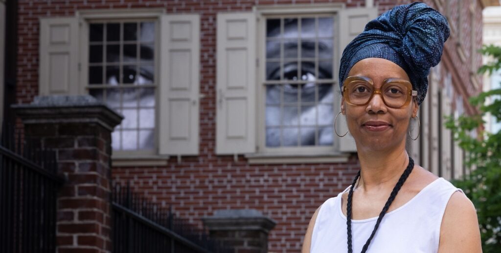 Sonya Clark, a Black artist, stands outside the Declaration House, a brick replica of the home where Thomas Jefferson wrote the "Declaration of Independence." In the paned windows are large video images of eyes belonging to the descendants of people Jefferson enslaved. Clakr is wearing her hair twisted into indigo fabric atop her head, large glasses, hoop earrings, a white tank and a black necklace.