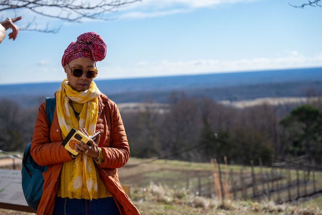 Sonya Clark, a Black woman wearing her hair twisted into a scarf-covered front-bun, looks through large sunglasses at she takes notes or reads in a small notebook in her hand. She is standing outside a Monticello, Thomas Jefferson's Charlottesville, VA estate, with his farmland in the background.