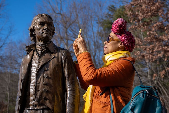 Monticello Site Visit and Portrait Sessions with Sonya Clark and Getting Word, Declaration House, Charlottesville, VA, 2024. Photo by AJ Mitchell for Monument Lab. Beside a bronze statue of Thomas Jefferson, a Black woman with her hair twisted in fabric into a front bun takes a photo with her phone (arms up), looking through large sunglasses and wearing an orange coat, yellow scarf with a turquoise leather backpack.