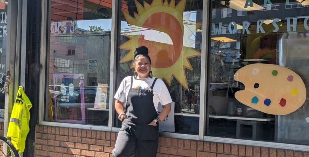 A Latina woman in grey overalls and a white shirt stands smiling in front of the cheerfully decorated glass windows of Sunshine House in Kensington, Philadelphia