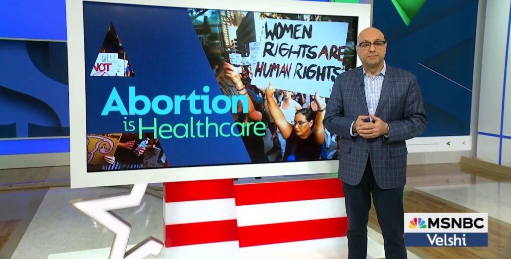 Show host Ali Velshi, a bald man with glasses in a grey suit and light blue shirt, stands beside a large monitor that reads Abortion is Healthcare and features women holding protest signs