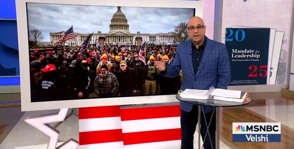 MSNBC host Ali Velshi, a bald man wearing glasses, a blue jacket, stands in front of a screen depicting the scene at the Capital on January 6, 2021. He is warning his TV audience about the dangers of Project 2025.