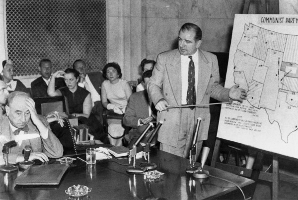 A black-and-white photo shows Senator Joe McCarthy (standing, pointing at a board) and attorney Joseph McCarthy (seated at a table). Behind these two suited White men is a partial row of seated people. 