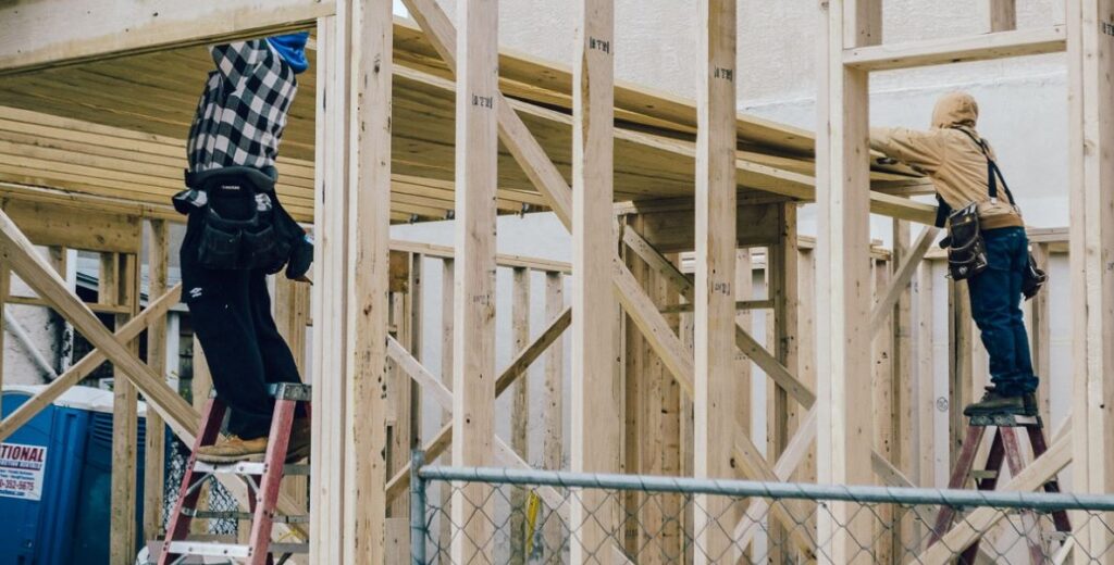 Construction workers on ladders assemble wood framing on a house under construction