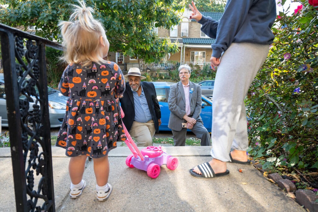 Maurice Sampson (left), an African American man with a wide-brim hat, khaki pants, navy suit jacket and blue collared shirt, stands beside Michael Kleiner, a white man with gray hair and gray mustach wearing a pink shirt, gray suit, buttons on his lapel and holding an iPhone, stand at the bottom of steps, facing a baby wearing a Halloween dress and pushing a plastic lawnmower and a person wearing slides, and sweats in Mount Airy in PA.