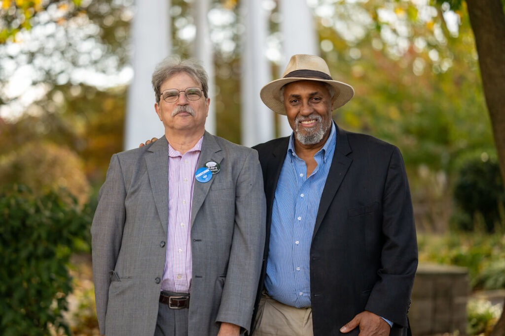 Michael Kleiner, a white man with gray hair and gray mustache wearing a pink shirt, gray suit, buttons on his lapel, stands beside Maurice Sampson (left), an African American man with a wide-brim hat, khaki pants, navy suit jacket and blue collared shirt. They are smiling.