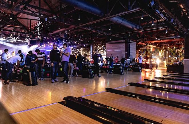 Inside of a bowling alley, with high ceilings and bowlers standing at the end of a long line of lanes.