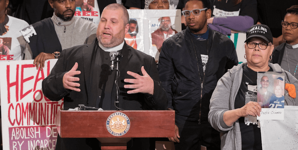 A large white man in a black suit and white collar, Reverend Christobal Kimmenez, stands at a wooden podium bearing the Pennsylvania coat of arms in front of a crowd advocating for criminal justice reform.