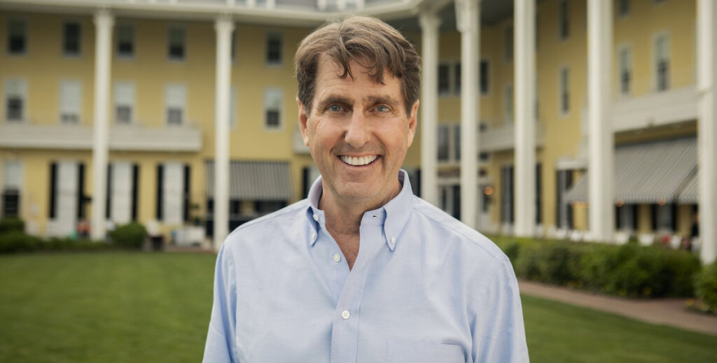 Curtis Bashaw, a 63-year-old White man with brown hair, blue eyes are large teeth, stands in front of historic Congress Hall, a yellow and columned large historic Victorian building facing the beach in Cape May, NJ that he converted into a luxury hotel.