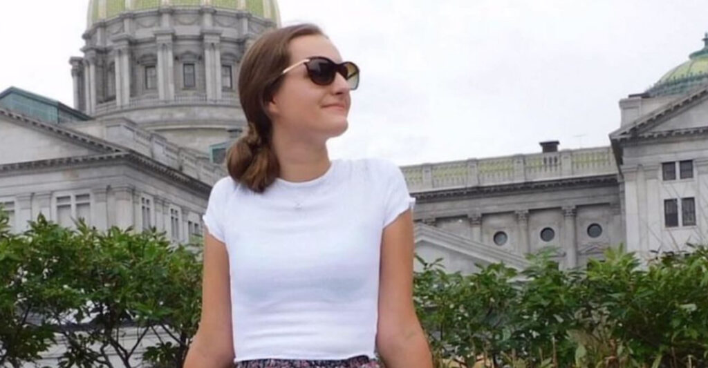 Erinda Sheno, a young white woman with brown hair pulled into a shoulder length ponytail, smiles and looks serenely to her left while seated outside in front of the capital building in Harrisburg, PA. She is wearing large sunglasses and a white short-sleeve t-shirt.